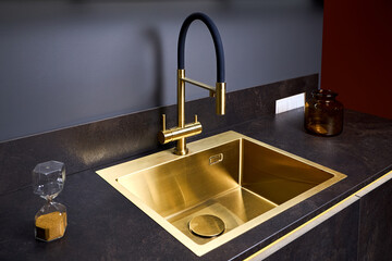 Luxurious interior square golden brass sink and faucet double tap mixer in contemporary modern design with stone marble stoneware countertop black and gold kitchen with sandwatch and glass pot.