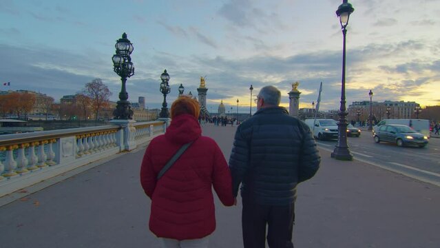 An elderly couple walks the streets of Paris along the famous bridge. Romantic relationships of the elderly. Journey of an elderly couple to the sights of Europe