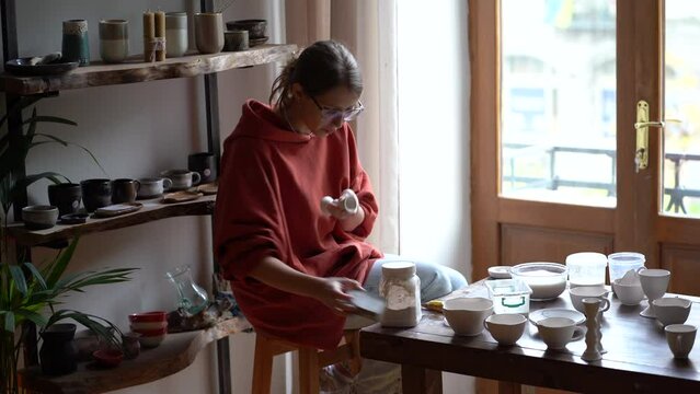 Concentrated attentive woman sits in workshop wiping items from kitchen utensils with rag and sponge. Self-sufficient casual businesswoman preparing porcelain items of own production for sale 