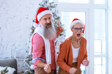 Fototapeta premium Christmas family grandmother and grandfather, elderly couple senior man and woman having fun near the Christmas tree, dancing and actively celebrating Christmas and New Year