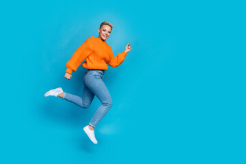 Fototapeta na wymiar Full length photo of cheerful excited lady wear orange sweatshirt jumping high running fast empty space isolated blue color background