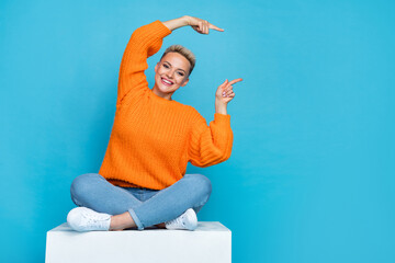 Photo of adorable pretty lady wear orange sweatshirt sitting platform pointing two finger empty space isolated blue color background - obrazy, fototapety, plakaty