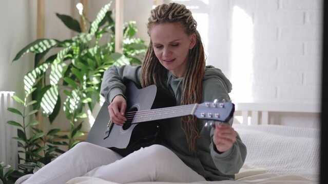 woman with dreadlocks on head singing song and playing acoustic guitar in sunny cottage bedroom, simple living and hobby relaxing