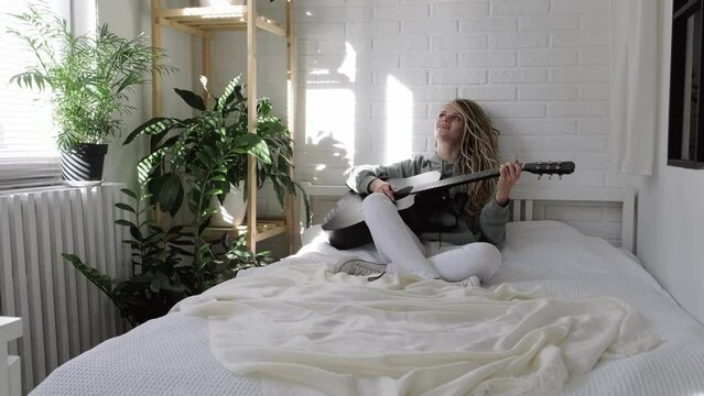 woman with dreadlocks on head singing song and playing acoustic guitar in sunny cottage bedroom, simple living and hobby relaxing