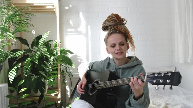 woman with dreadlocks on head singing song and playing acoustic guitar in sunny cottage bedroom, simple living and hobby relaxing