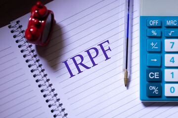 A sigla IRPF para Imposto sobre a Renda das Pessoas Físicas escrita em um caderno. Relógio e Calculadora na composição. Imposto de Renda no Brasil.  - obrazy, fototapety, plakaty