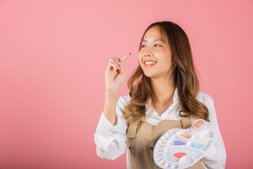 Asian beautiful young woman artist holding brush and paint palette, Happy female painting using paintbrush and palette with colors, studio shot isolated on pink background, Paintings and art equipment