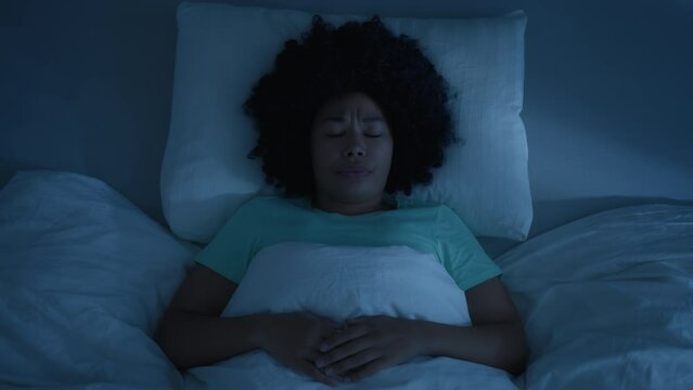 Young female laying in bed sleeping, suddenly woken up by nightmare, screaming with wide opened eyes 4K. Beautiful African American woman waking up through a nightmare on dark night in blue moonlight