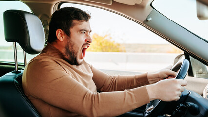 A young male driver screams loudly while sitting at the wheel and looks somewhere on the road. A...