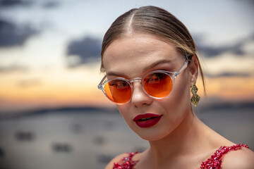 Fototapeta na wymiar Beautiful female model wearing colorful glasses at sunset. Outdoors romantic portrait of attractive blonde woman with makeup and glasses posing. Istanbul archipelago (Princess Islands) skyline.