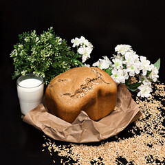 A loaf of freshly baked homemade rye bread lies on craft paper against the background of scattered grains and flowers, a glass of milk, simple and healthy food