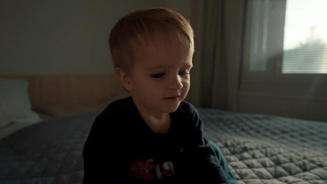 Curious little boy points finger to pictures in book sitting on bed against bright window. Toddler spends morning in bedroom closeup slow motion