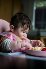 An eight-month-old baby, who is eating fruit for the first time, examines the fruit on his plate. Close up