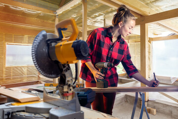 Woman repairman. Girl carpenter inside house under construction. Carpenter makes mark with pencil on board. Woman carpenter with electric screwdriver. Girl builds house with her own hands