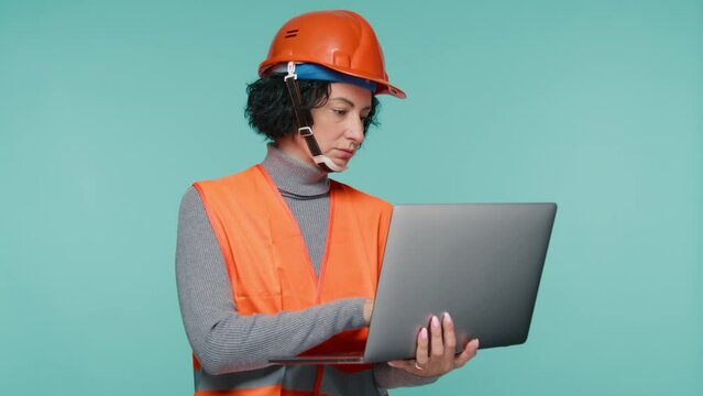 Female civil engineer reviewing future of real estate development planning. Wearing Safety helmet and vest, city construction 4K. Young female building architect use laptop on blue studio background