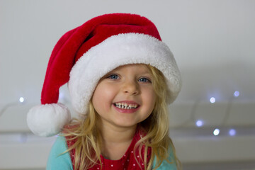 Little blond girl in red Santa hat and Christmas decoration on background looking at the camera and smiling,portrait of kid in New year costume,winter holidays image.