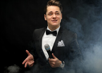 Stylish young man in a tuxedo holding a microphone, posing against a dark background with smoke,...