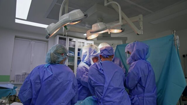 Medical team standing around the operational table under the bright lamps. Nurse passes the instruments to the surgeons.