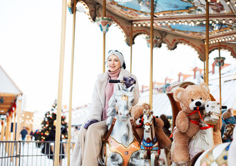 Fototapeta na wymiar Cheerful young woman in a faux fur coat and mittens rides a carousel and has fun