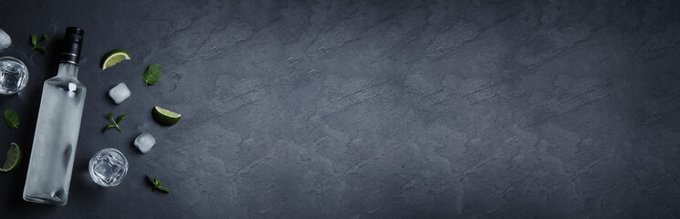 Bottle of vodka, shot glasses, lime, mint and ice on black table, flat lay. Space for text. Banner design