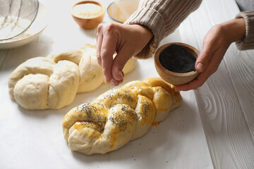 Woman adding poppy seeds onto braided dough at white wooden table in kitchen, closeup. Cooking...