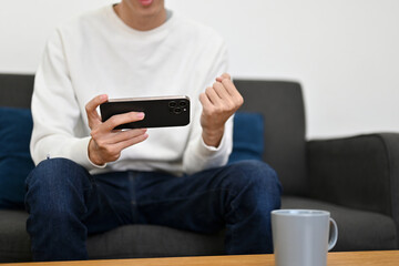 A young man sits on sofa showing clenched fist while playing a mobile game or watching football