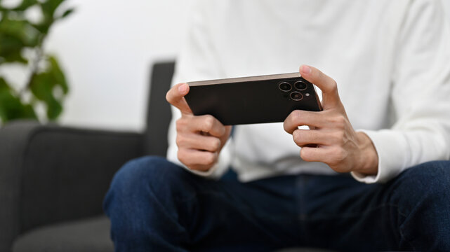 A young man sits on sofa playing a mobile game, watching movie on his smartphone. cropped