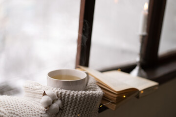 Cozy winter still life. Cup of hot tea and an open book with a warm sweater on a vintage wooden windowsill. Cozy home concept. Sweet home.