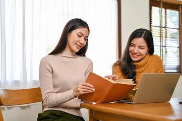 Two happy Asian female college friends research some information in the book