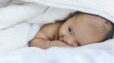Cute adorable baby boy hidden in white blanket on the bed, laying prone and looking to the side while smiling.