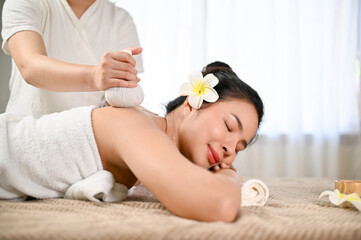 Obraz na płótnie Canvas Asian woman lying on spa massage table, eyes closed, getting herbal ball stamp on her back