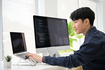 Smart young Asian male programmer coding at his desk, using laptop and computer.