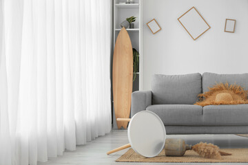 Interior of messy living room with grey sofa, shelving unit and surfboard