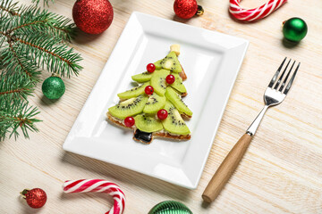 Plate of tasty pancake in shape of Christmas tree with fruits, fir branches and decorations on wooden background