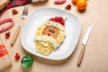 Plate with Santa Claus made of pancake, fruits, whipped cream and Christmas decor on beige table