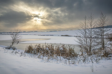 Winter snowy landscape with frozen pond and sun rays
