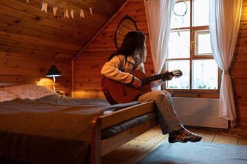 Melancholic lonely woman sits on bed with guitar in hands and looks out window admiring beautiful...
