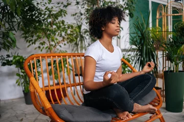 Fotobehang Mindful barefoot black young woman sitting with closed eyes on sofa in lotus pose, practicing yoga meditation at home, relaxing and breathing deep for mental health in room with biophilia design. © dikushin