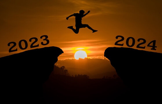 A young man jump between 2023 and 2024 years over the sun and through on the gap of hill silhouette evening colorful sky. happy new year 2023.