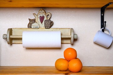 Three large ripe orange oranges lie on a wooden table in the farmhouse. On the wall hangs a ceramic mug and a roll of paper towels.