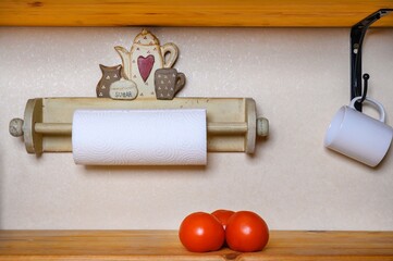 Three large, ripe red tomatoes lie on a wooden table in a farmhouse. On the wall hangs a ceramic mug and a roll of paper towels.