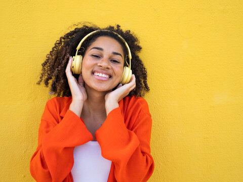 Young african american teenage girl listening to music happily with headphones over yellow wall - Powered by Adobe