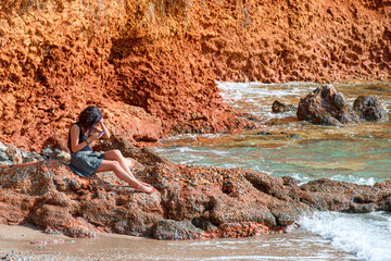 A young woman is sitting and using her smartphone at the rocky shore on the background of the sea.