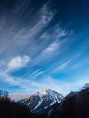 青い青空一面に白い筋雲、谷間の奥には雪を被った山、山頂に日差しが当たる。