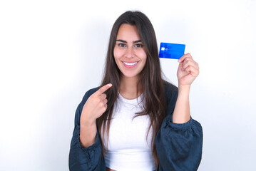 Young brunette woman wearing casual clothes standing over white studio background holding a credit card and pointing with forefinger at it. 