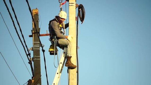 Skilled electrician in helmet fixes wires standing on ladder near high pole against blue sky on summer day backside view. Electrical service and mounting on the pole. Slow motion