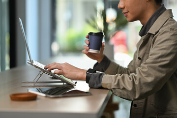 Side view of young man drinking coffee from paper cup and working online on laptop computer