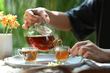 Cropped view of young man pouring traditional tea from transparent teapot into a glass cup