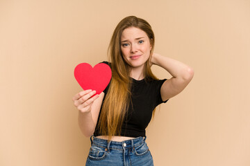 Young redhead woman holding a paper red heart cut out isolated touching back of head, thinking and making a choice.