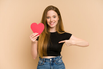 Young redhead woman holding a paper red heart cut out isolated person pointing by hand to a shirt copy space, proud and confident